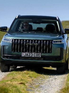 Front view of a green Ineos SUV driving on a grassy path with a visible licence plate.