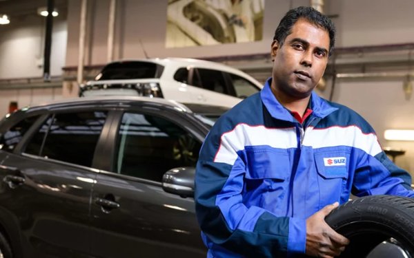 Man in a blue uniform holding a tyre near a car in a garage.